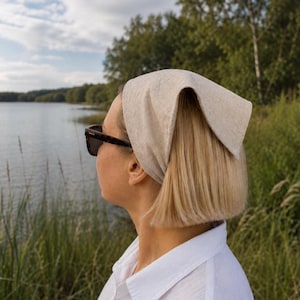 May include: A light beige headscarf tied in a knot, worn on the head. The headscarf is made of a textured fabric. The person is wearing dark sunglasses and a white collared shirt. The background includes a lake and trees.