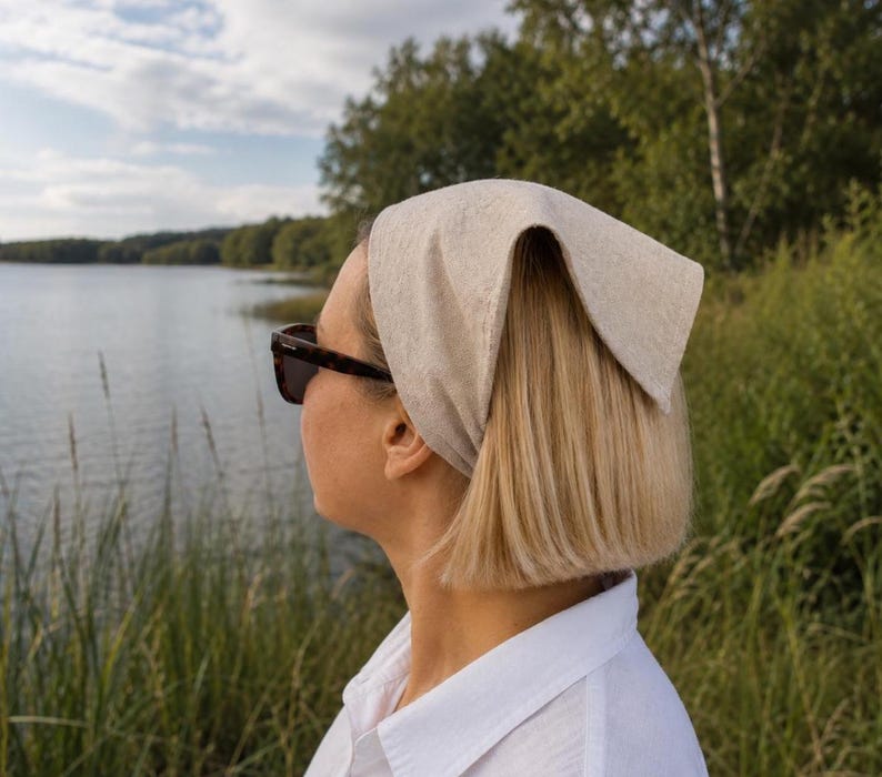 May include: A light beige headscarf tied in a knot, worn on the head. The headscarf is made of a textured fabric. The person is wearing dark sunglasses and a white collared shirt. The background includes a lake and trees.