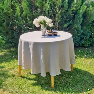 May include: A round, light gray linen tablecloth draped over a wooden table with four legs. A white vase with white flowers sits on the table, along with three black and gold teacups.