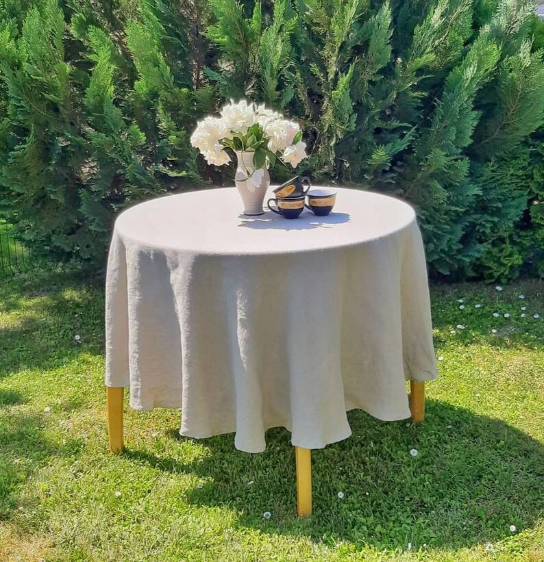May include: A round, light gray linen tablecloth draped over a wooden table with four legs. A white vase with white flowers sits on the table, along with three black and gold teacups.