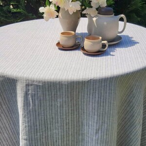 May include: A white linen tablecloth with gray stripes, a teapot, two teacups, and a bouquet of white peonies in a ceramic vase.