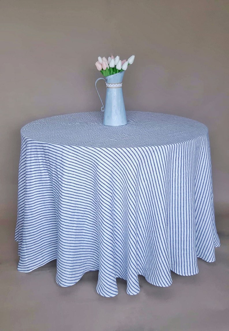 May include: A white tablecloth with blue diagonal stripes, featuring a blue metal pitcher with a bouquet of pink and white tulips.
