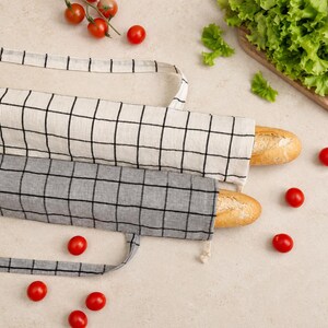 May include: Two long, rectangular fabric bread bags with handles, each containing a baguette. One bag is off-white with a black grid pattern, and the other is gray with a black grid pattern. The bags are made of a woven textile material.