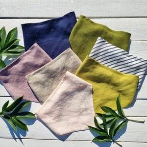 May include: A collection of folded linen face masks in various colors, including navy blue, olive green, and pale pink. One mask features blue and white stripes. The masks are arranged on a white wooden surface, with green leaves in the corners.