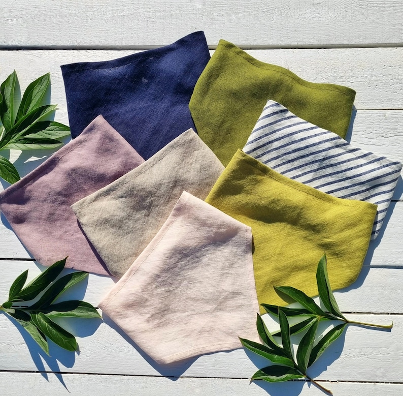 May include: A collection of folded linen face masks in various colors, including navy blue, olive green, and pale pink. One mask features blue and white stripes. The masks are arranged on a white wooden surface, with green leaves in the corners.