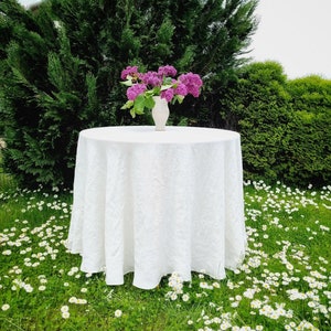 Può includere: Una tovaglia di lino bianca con un delicato motivo floreale, posta su un prato verde con margherite bianche. Un vaso di ceramica bianco con un bouquet di fiori di lillà viola è posto al centro del tavolo.