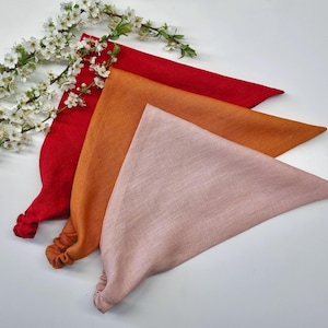 Könnte beinhalten: Drei dreieckige Kopftücher mit elastischen Bändern in den Farben Rot, gebranntes Orange und zartem Rosa. Der Stoff weist eine sichtbare Webstruktur auf und die Artikel sind in einem überlappenden Stapel angeordnet.