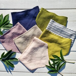 May include: A collection of folded linen face masks in various colors, including navy blue, olive green, and pale pink. One mask features blue and white stripes. The masks are arranged on a white wooden surface, with green leaves in the corners.
