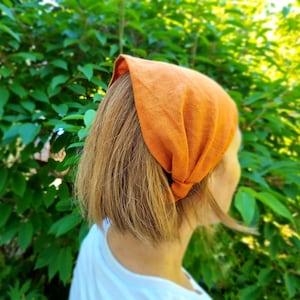 May include: An orange linen headband is worn on the head. The headband is knotted at the back and covers the top of the head. The person is wearing a white shirt. The background is a lush green bush.