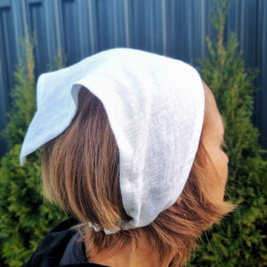 May include: A white linen head covering, possibly a bonnet or scarf, is worn on the head. The fabric appears soft and slightly textured. The background includes green foliage and a dark fence.