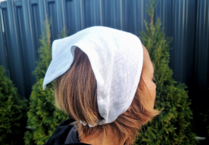 May include: A white linen head covering, possibly a bonnet or scarf, is worn on the head. The fabric appears soft and slightly textured. The background includes green foliage and a dark fence.