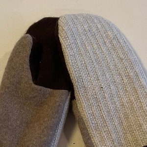 Op de afbeelding: Een paar bruine, beige en witte gebreide wanten met een donkergrijze manchet. De wanten zijn gemaakt van een zacht, warm materiaal en perfect om je handen warm te houden in de winter.
