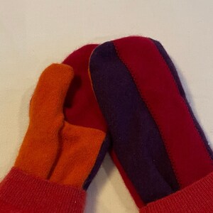 Op de afbeelding: Een paar gebreide wanten met een gestreept ontwerp. Eén want is oranje, rood en paars, terwijl de andere rood en paars is. De wanten hebben rode manchetten.