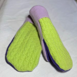 Op de afbeelding: Een paar groene en paarse gebreide wanten met een kabelbreipatroon. De wanten hebben een lichtpaarse manchet en een donkerpaarse rand.