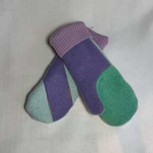 Op de afbeelding: Een paar kleurrijke wanten in tinten paars, groen en lichtblauw. De wanten hebben een geribbelde lavendelkleurige manchet en zijn gemaakt van een zacht, getextureerd materiaal. Het ontwerp heeft color-blocked secties en decoratieve stiksels.
