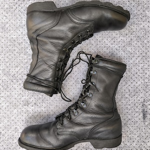 Könnte beinhalten: Ein Paar schwarze Leder-Kampfstiefel mit hohem Schaft, abgerundeten Zehenkappen und dicken Gummisohlen. Die Stiefel verfügen über schwarze Schnürsenkel, die durch Metallösen und Schnürhaken geführt werden, sowie einen gepolsterten Schaftrand.