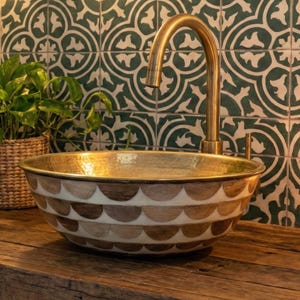 May include: A bathroom sink with a gold interior and a decorative exterior featuring a pattern of brown and white half-circles. The sink is set on a rustic wooden vanity. A brass faucet and a green and white patterned tile wall are in the background.