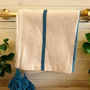 Könnte beinhalten: Ein cremefarbenes Handtuch mit einem türkisfarbenen Streifen und Besatz hängt an einem Messing-Handtuchhalter. Eine türkisfarbene Quaste hängt an der Unterseite des Handtuchs. Das Handtuch wird vor einem hellen Holzhintergrund mit grünen Pflanzen auf beiden Seiten präsentiert.