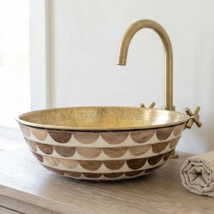 May include: A decorative bathroom sink with a gold interior and a patterned exterior in shades of brown and cream. A gold faucet and rolled beige towel sit on a wooden cabinet.