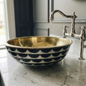May include: A gold-colored, round bathroom sink with a black and white scalloped pattern. The sink is set on a white marble countertop with a silver faucet. The sink is a decorative element in a bathroom.