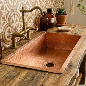 May include: A bathroom with a copper sink set into a reclaimed wood vanity. The sink has a brass tap and is surrounded by amber glass bottles. A woven basket holds rolled towels, and a hanging plant adds greenery. The tiles have a patterned design.