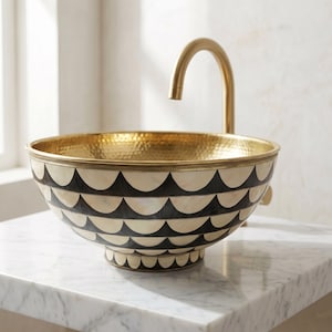 May include: A round vessel sink featuring a hammered gold interior and an exterior decorated with a repeating black and cream scalloped pattern. A curved gold faucet extends over the basin, which is mounted on a white marble surface.