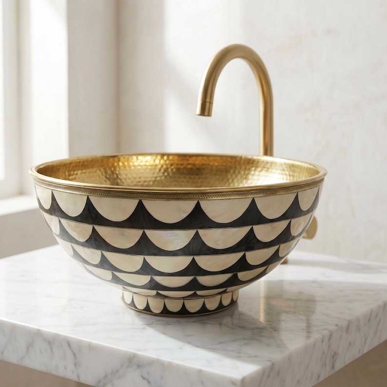 May include: A round vessel sink featuring a hammered gold interior and an exterior decorated with a repeating black and cream scalloped pattern. A curved gold faucet extends over the basin, which is mounted on a white marble surface.