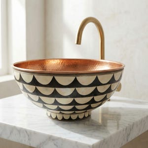 May include: A decorative copper sink with a hammered interior and a patterned exterior. The sink features a black and cream scallop design and a gold faucet. The sink is a unique bathroom fixture.