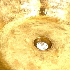May include: A round, metallic gold-colored sink basin featuring a distinct hammered texture across its entire interior surface. The basin has a smooth, circular drain opening located in the center, reflecting light across the polished metal finish.