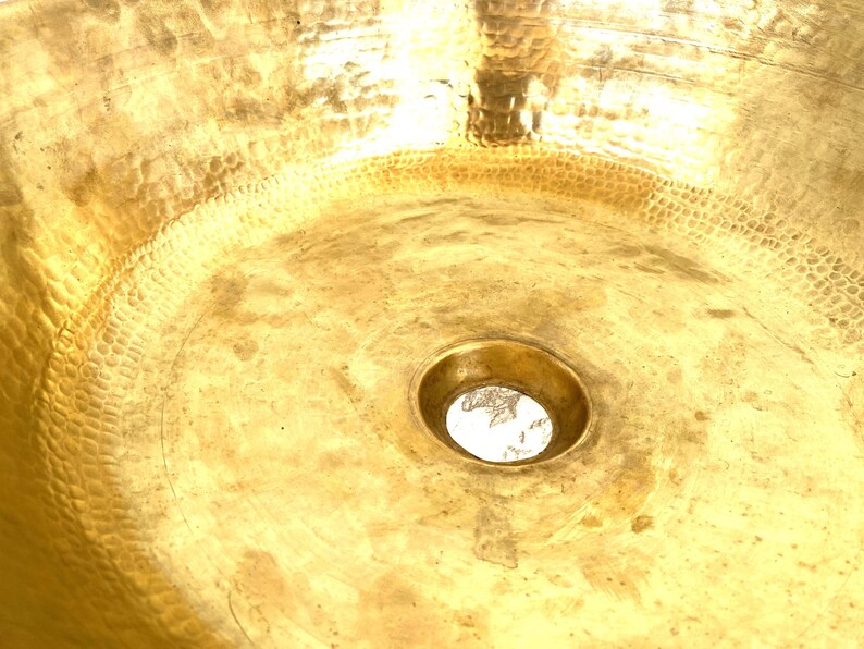 May include: A round, metallic gold-colored sink basin featuring a distinct hammered texture across its entire interior surface. The basin has a smooth, circular drain opening located in the center, reflecting light across the polished metal finish.