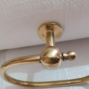 May include: A brass towel ring holder with a circular design. The holder has a round mounting plate and a curved arm that forms the ring. The finish is a shiny gold color, and the design is simple and elegant.
