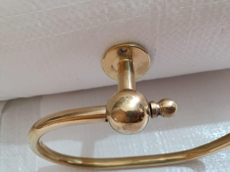 May include: A brass towel ring holder with a circular design. The holder has a round mounting plate and a curved arm that forms the ring. The finish is a shiny gold color, and the design is simple and elegant.