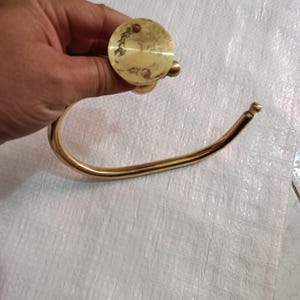 May include: A vintage brass towel ring with a circular mounting plate and a curved, open ring. The brass has a warm, golden hue with signs of age and wear. The design is simple and functional, suitable for a bathroom or kitchen.