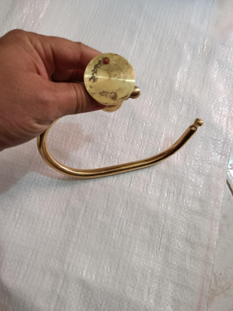 May include: A vintage brass towel ring with a circular mounting plate and a curved, open ring. The brass has a warm, golden hue with signs of age and wear. The design is simple and functional, suitable for a bathroom or kitchen.
