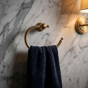 May include: A gold-toned towel ring holds a navy blue towel against a marble wall. A matching wall sconce with a lampshade is to the right. The bathroom fixtures have a classic, elegant design.