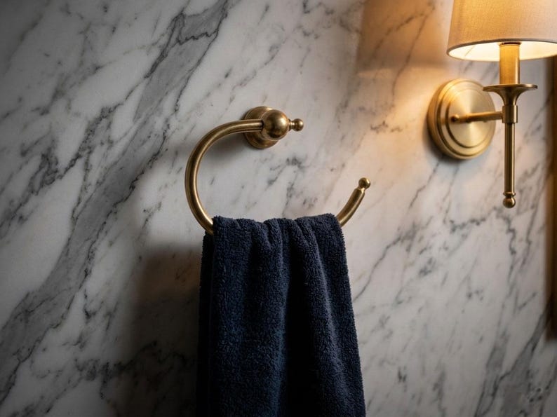 May include: A gold-toned towel ring holds a navy blue towel against a marble wall. A matching wall sconce with a lampshade is to the right. The bathroom fixtures have a classic, elegant design.