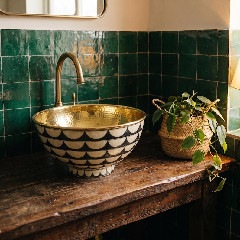 May include: A round vessel sink featuring a hammered gold interior and a black and white scalloped pattern on the exterior. The sink is paired with a curved gold faucet and sits on a dark wooden surface next to a green plant in a woven basket.