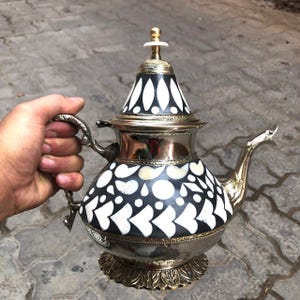 Puede incluir: Una tetera de plata con un patrón geométrico en blanco y negro. La tetera tiene un cuerpo redondo, un pico, una manija y una tapa con un pomo. La tetera está decorada con un patrón de círculos blancos y formas negras.