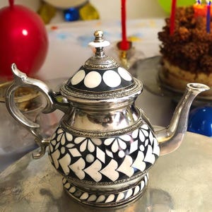 May include: A silver teapot with a black and white geometric pattern. The teapot has a round body and a spout on the side. The teapot is decorated with white circles and a white floral design.