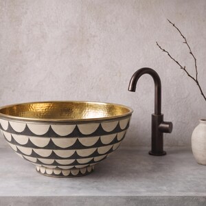 May include: A round vessel sink featuring a hammered gold interior and an exterior decorated with a repeating black and white scalloped pattern. The sink is paired with a dark bronze gooseneck faucet and a small beige vase with a bare twig.