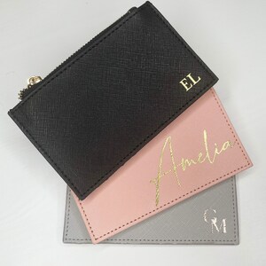 May include: Three personalised card holders in black, pink, and grey. The black card holder has a zip and gold initials "EL". The pink card holder has the name "Amelia" in gold script. The grey card holder has silver initials "GM".