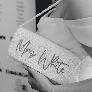 May include: A white beaded clutch bag with the inscription "Mrs White" in cursive. The rectangular bag has a beaded strap and is held by a person wearing a white dress. A silver bracelet is visible.