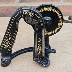May include: An antique black and gold sewing machine component. The metal piece features ornate gold detailing and a hand crank. The design includes a curved handle and a circular wheel, showcasing a vintage aesthetic.