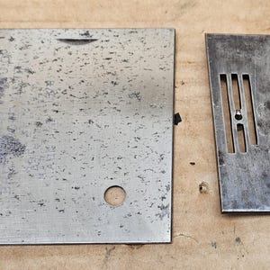 May include: Two vintage sewing machine parts. One is a rectangular needle plate with a hole and a slot. The other is a smaller, rectangular throat plate with three needle slots and a hole. Both are made of metal.
