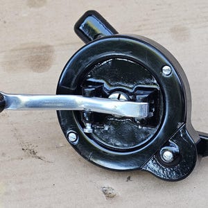May include: A vintage manual hand-cranked tool featuring a glossy black circular metal housing, a silver-toned metal crank arm, and a dark brown wooden handle. The mechanism includes exposed screws and a small metal attachment point on the side.
