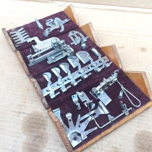 May include: A wooden box with a burgundy velvet lining containing a collection of vintage sewing machine feet and attachments. The box is open and the feet are arranged in rows.