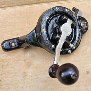 May include: A vintage black metal hand-cranked drill with a silver-colored metal handle and a dark brown spherical knob. The tool features a circular gear housing, a mounting arm with a circular hole, and visible signs of wear on the black paint.