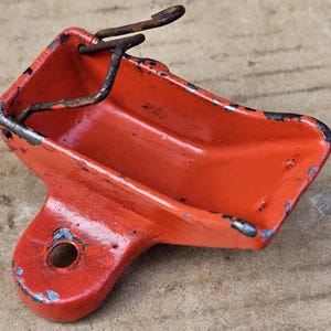 Op de afbeelding: Oranje metalen batterijhouder met een veerbelaste deksel. De houder heeft een gat aan de zijkant en een roestige metalen veer.
