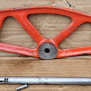 May include: An orange metal tool with a long, thin arm and a circular opening in the centre. The tool has a smaller, cylindrical piece attached to the arm. A separate, silver metal rod is also pictured.