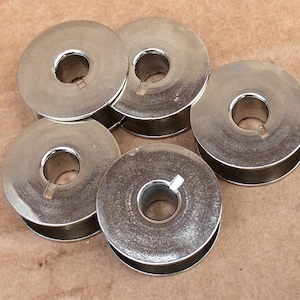 May include: Five circular metal sewing machine bobbins with a polished silver finish. Each bobbin features a central hole and a small notch on the inner rim, designed for holding thread securely during the sewing process for various textile projects.